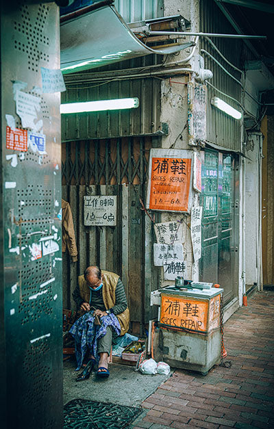 A metal print featuring a chromoluxe finish, depicting a vibrant cityscape at night with illuminated store fronts and a person sitting beside a shoebeat repair stand.