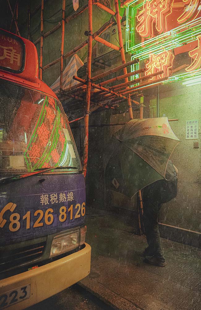 A photograph capturing urban street scenes in Hong Kong, featuring a bus, neon signs, and a person with an umbrella.