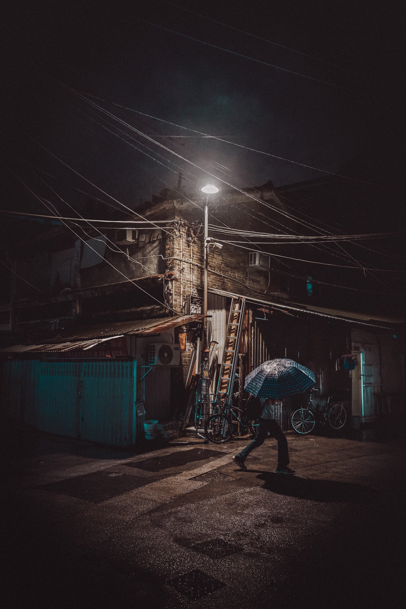 A person with an umbrella walking in a village scene at night. The image has a dark tone with a street lamp providing light and power lines in the background.