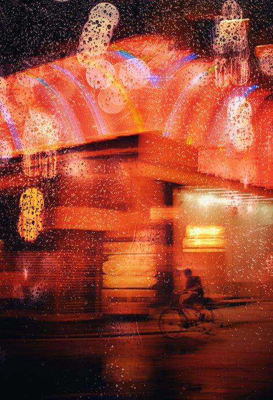 An abstract, artistic photograph of a rainy night street scene with blurred lights, warm orange and red glow, and a small cyclist near the bottom right.