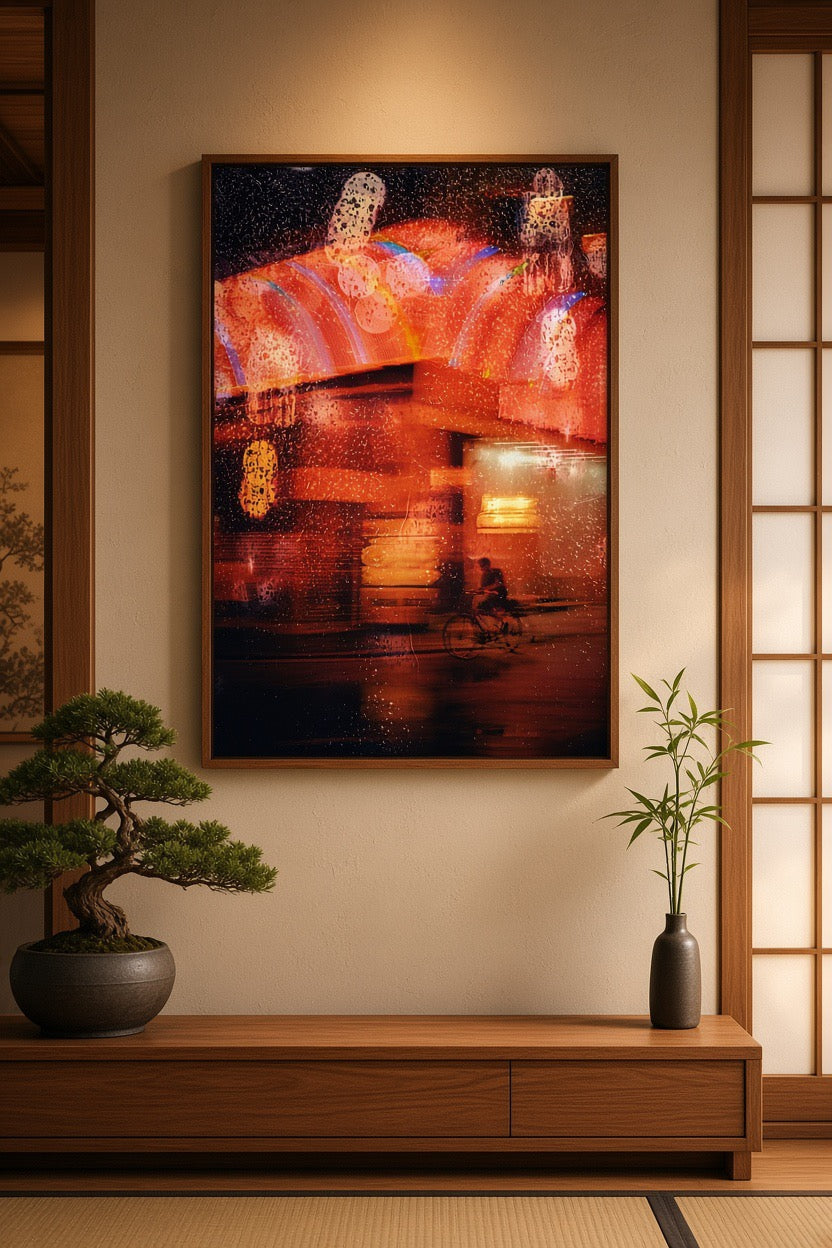 A framed vertical art print depicting a blurred, warm-toned street scene with glowing lanterns and reflections, displayed on a beige wall above a wooden console table in a styled interior with a bonsai tree and a bamboo plant.
